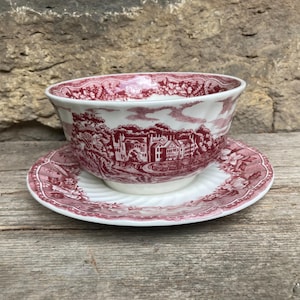 May include: A white ceramic bowl and matching saucer set featuring a detailed red transferware pattern. The design depicts a landscape with a large house, trees, and clouds, complemented by a decorative floral border along the rim of both pieces.