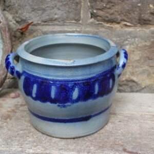 Pode incluir: Um pote de cerâmica azul e branco com duas alças. O pote tem um design azul e branco ao redor do meio.