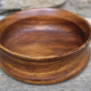 Puede incluir: Un cuenco redondo de madera de color marr&oacute;n c&aacute;lido y rico. El cuenco tiene una base ligeramente elevada y un interior liso y pulido. La veta de la madera es visible, lo que a&ntilde;ade a su est&eacute;tica natural. Adecuado para servir alimentos o como objeto decorativo.