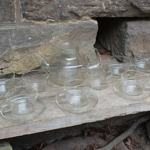 Op de afbeelding: Een set van heldere glazen theekopjes en schoteltjes met een bijpassende glazen theepot. De kopjes en schoteltjes zijn gerangschikt op een houten oppervlak.