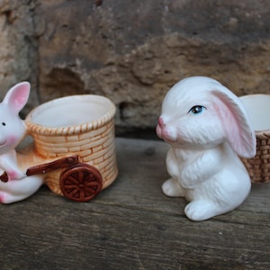 Könnte beinhalten: Zwei weiße Keramik-Hasenfiguren. Ein Hase zieht einen kleinen braunen Weidenkorb auf Rädern. Der andere Hase hält einen kleinen braunen Weidenkorb.