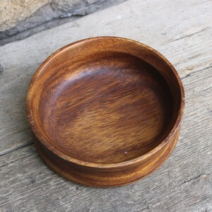 Puede incluir: Un cuenco redondo de madera, de color marr&oacute;n oscuro con vetas visibles. El cuenco tiene un dise&ntilde;o sencillo y cl&aacute;sico, ideal para servir alimentos o como objeto decorativo. Se encuentra sobre una superficie de madera envejecida.