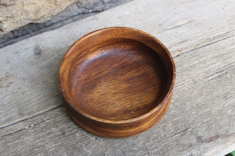 Puede incluir: Un cuenco redondo de madera, de color marr&oacute;n oscuro con vetas visibles. El cuenco tiene un dise&ntilde;o sencillo y cl&aacute;sico, ideal para servir alimentos o como objeto decorativo. Se encuentra sobre una superficie de madera envejecida.