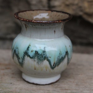 Puede incluir: Un pequeño jarrón de cerámica blanca con un borde marrón y un patrón de esmalte verde y blanco. El jarrón tiene un cuerpo redondeado y un labio acampanado.