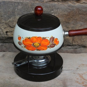 Può includere: Un pentolino per fondue vintage bianco con un coperchio e un manico marroni. Il pentolino presenta un motivo floreale con fiori arancioni e marroni. Il pentolino è posto su un supporto nero con un bruciatore.