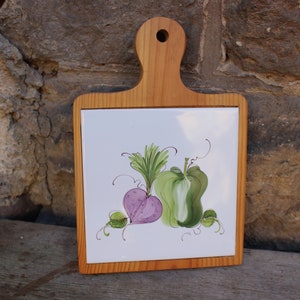 May include: A wooden cutting board with a white ceramic tile inset. The tile features a hand-painted design of a purple beet and a green pepper.