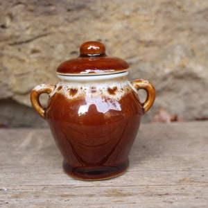 Könnte beinhalten: Eine kleine braune Keramikdose mit Deckel. Die Dose hat eine weiße Glasur mit braunen Flecken. Die Dose hat zwei Griffe an den Seiten.