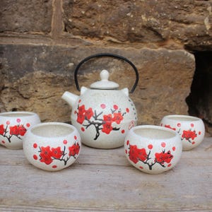 May include: A ceramic tea set featuring a cream-colored teapot and four matching cups. Each piece is adorned with a red floral design and black stems. The teapot has a black handle and a small lid. The set is displayed on a wooden surface.