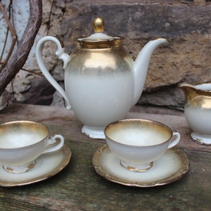 May include: A vintage tea set with a white teapot, creamer, and two cups and saucers. The set features a gold trim and a subtle ombre effect.