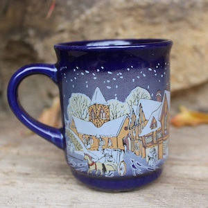 Puede incluir: Una taza de cerámica azul con una escena de invierno de un pueblo nevado con un carruaje tirado por caballos. La taza tiene un borde y un asa blancos.