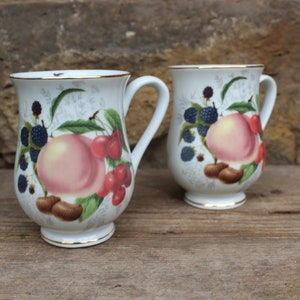 以下が含まれることがあります： 金色の縁取りのある白いセラミック製のマグカップが2つ。マグカップには、桃、サクランボ、ブラックベリーが描かれた花柄のデザインが施されています。