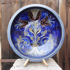 May include: A large blue ceramic bowl with a decorative floral design in gold and brown. The bowl has a raised rim and is sitting on a wooden stand.