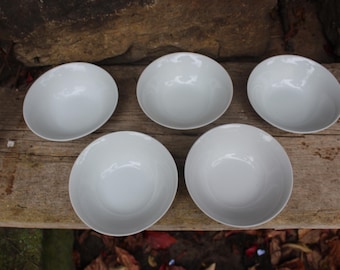 May include: Five white ceramic bowls arranged on a weathered wooden surface. The bowls are round with a smooth, glossy finish. The bowls are empty and ready for use. The background includes a rock and some foliage.