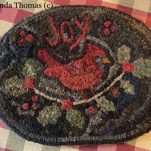 May include: Oval-shaped, hand-stitched rug with a cardinal bird design. The rug features the word "JOY" in red, a red cardinal, green leaves, and red berries on a dark background. The rug is approximately 12 inches in diameter.