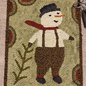 May include: A rectangular rug hooking project on burlap featuring a snowman wearing a dark hat, a red scarf, and brown overalls with suspenders. The background is sage green with circular patterns and a pine branch. Text reads: Jolly hooked by Rhonda Thomas.