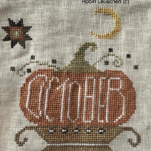 Op de afbeelding: Kruissteekontwerp met een pompoen met het woord "OCTOBER" in wit geborduurd. De pompoen is in tinten oranje en bruin, met een groene steel. Een halve maan en een ster zijn ook geborduurd.