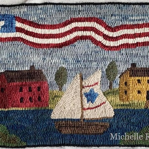 May include: Hand-hooked rug depicting an American flag waving above a coastal scene. The rug features a red house, a yellow house, a sailboat, and a lighthouse. The artist's name, Michelle Robinson, is in the bottom right corner.