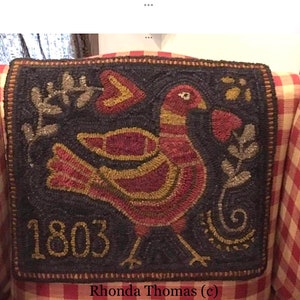 May include: A dark brown rug with a red and gold bird design and the year 1803 embroidered in gold. The rug is on a red and white checkered chair.