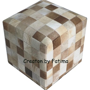 May include: A square ottoman with a patchwork design made of different shades of brown and beige cowhide. The ottoman is made of leather and has a square shape. The ottoman is made by Creation by Fatima.