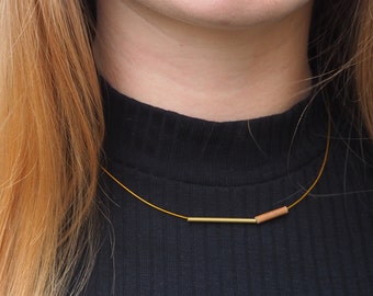 May include: A delicate gold-colored necklace with a thin chain. The necklace features a horizontal gold bar and a small, cylindrical, reddish-brown bead. The necklace is worn against a black top.