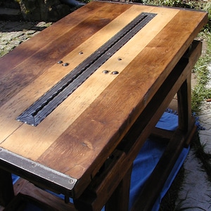 Könnte beinhalten: Ein rustikaler Holztisch mit einem Metallstreifen in der Mitte. Der Tisch ist aus dunklem und hellem Holz gefertigt und hat eine natürliche Oberfläche. Der Tisch steht auf einer blauen Decke.