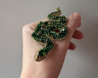 May include: A green snake brooch made of beads and crystals. The snake is coiled and has a green gemstone for an eye.