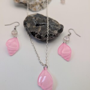May include: A pink jewellery set comprising a necklace and earrings. The necklace has a silver chain with a pink pendant. The earrings feature pink shell-shaped pendants and small clear beads. The set is displayed on a white surface.