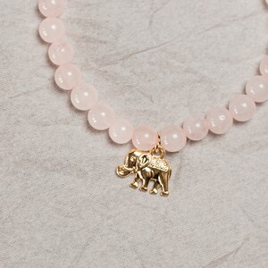 May include: A rose quartz beaded bracelet with a gold elephant charm. The bracelet features round, light pink beads and a small gold accent bead. The elephant charm is detailed with etched designs.