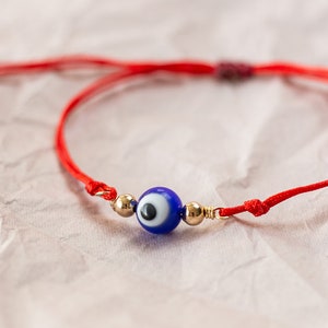 Puede incluir: Una pulsera de hilo rojo con una cuenta de ojo turco azul y detalles dorados. La pulsera está anudada y se muestra sobre una superficie rosa claro texturizada.