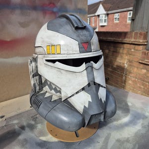 Può includere: Un casco dettagliato dipinto in bianco, grigio e nero. Presenta una visiera, un triangolo rosso e tre rettangoli gialli. Il design include motivi angolari. Il casco è posto su una base circolare marrone.
