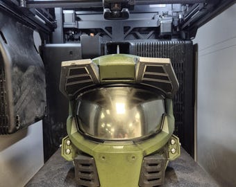 Op de afbeelding: Een groene Master Chief helm met een donker reflecterend vizier en een prominente voorhoofdsplaat, geplaatst in een 3D-printer. De helm heeft zwarte ventilatieroosters aan de voor- en zijkanten, met een textuur op het olijfgroene oppervlak.