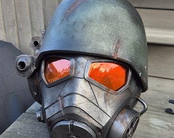 May include: A distressed, grey metallic helmet with orange lenses. The helmet has a gas mask-style respirator, side-mounted cylinders, and a textured surface, evoking a post-apocalyptic or sci-fi aesthetic.