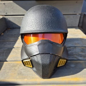 以下が含まれることがあります： オレンジ色のバイザーと金色のアクセントが付いた黒いヘルメット。ヘルメットはテクスチャ加工された表面と、前面に幾何学模様のデザインがあります。バイザーは光を反射し、明るいオレンジ色の輝きを生み出します。