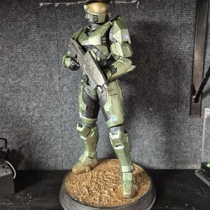 Könnte beinhalten: Detaillierte Figur eines grün-schwarzen gepanzerten Soldaten, der ein Gewehr hält. Die Figur steht auf einem kreisförmigen Sockel mit einer strukturierten, braunen Oberfläche. Der Helm des Soldaten hat ein goldenes Visier.
