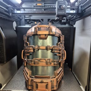May include: A copper-colored, futuristic helmet with a clear visor, likely a cosplay or prop item. The helmet is detailed with various panels and rivets, and is sitting on a 3D printer bed.