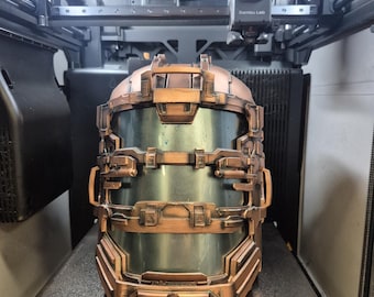 May include: A copper-colored, futuristic helmet with a clear visor, likely a cosplay or prop item. The helmet is detailed with various panels and rivets, and is sitting on a 3D printer bed.