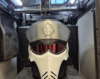 May include: A 3D printed helmet featuring a metallic silver forehead plate with a circular emblem, a dark red visor, and a white lower face mask with black accents. The helmet is positioned inside a 3D printer with a 320 x 340 mm build plate.