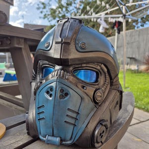 Puede incluir: Casco futurista azul y bronce con lentes azules reflectantes. El casco presenta un diseño detallado con ventilaciones, remaches y una máscara facial. Probablemente para cosplay o exhibición.