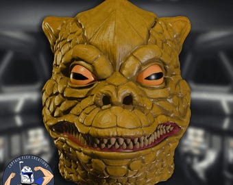May include: A detailed, mustard-colored mask resembling a reptilian creature. The mask features orange eyes, a wide, toothy grin with red gums, and textured skin. The mask is likely for cosplay or costume use.