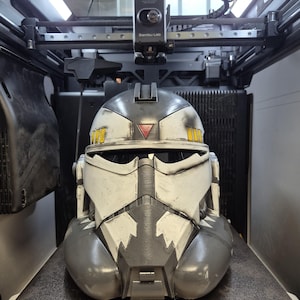 May include: A white and gray clone trooper helmet with weathered detailing, featuring a red triangle on the forehead, yellow rectangular accents on the sides, and a jagged black pattern around the mouth, resting inside a 3D printer build plate.