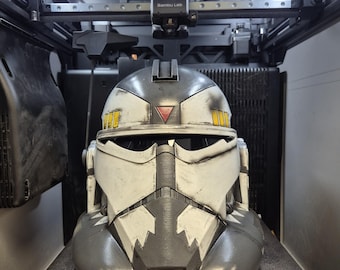 May include: A white and grey clone trooper helmet with weathered detailing, featuring a red triangle on the forehead, yellow rectangular accents on the sides, and a jagged black pattern around the mouth, resting inside a 3D printer build plate.