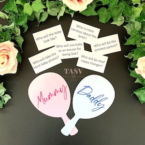 May include: A set of two pink and white paddle-shaped game pieces with the words "Mummy" and "Daddy" written on them. The paddles are surrounded by white cards with questions about a new baby, such as "Who will the baby look like?" and "Who will be the strictest parent?"