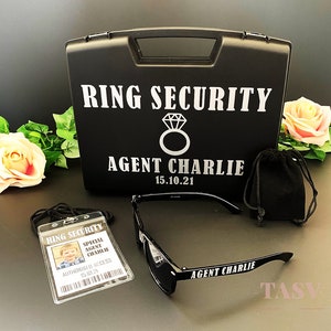 May include: Black ring security case with a diamond ring icon and the text "RING SECURITY" and "AGENT CHARLIE 15.10.21".  A black ID badge with the text "RING SECURITY" and "SPECIAL AGENT CHARLIE" and a black pouch.