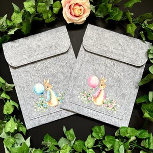 May include: Two grey felt document sleeves with flap closures. Each features a printed illustration of a rabbit holding a balloon amidst flowers. One sleeve reads FINNIAN'S LITTLE RED BOOK, the other reads CORA'S LITTLE RED BOOK in white text.