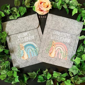 May include: Two grey felt book covers with a bunny and rainbow design. One cover says "William's Little Red Book" with a blue rainbow and the other says "Charlotte's Little Red Book" with a pink rainbow.