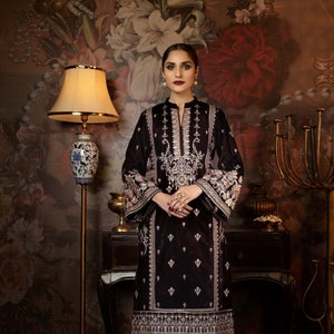 May include: A woman wearing a black velvet kurta with silver embroidery and matching trousers. The kurta has a high neckline and long sleeves. The woman is standing in front of a floral wallpaper.
