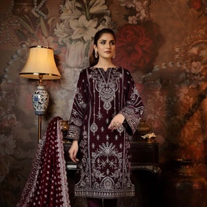 May include: A maroon velvet dress with silver embroidery and a matching maroon velvet dupatta. The dress has a round neckline and long sleeves. The dupatta is decorated with a silver border. The model is wearing the dress with maroon pants and beige heels.