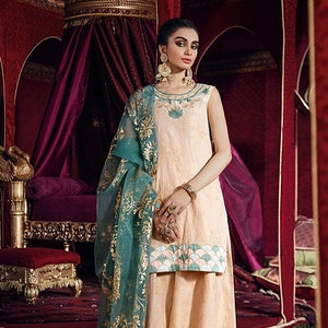 May include: A woman wearing a peach-colored sleeveless dress with gold embroidery and a teal dupatta with gold accents. The dress has a scalloped hemline with a pink and teal design. The woman is standing in a room with a red velvet canopy bed.