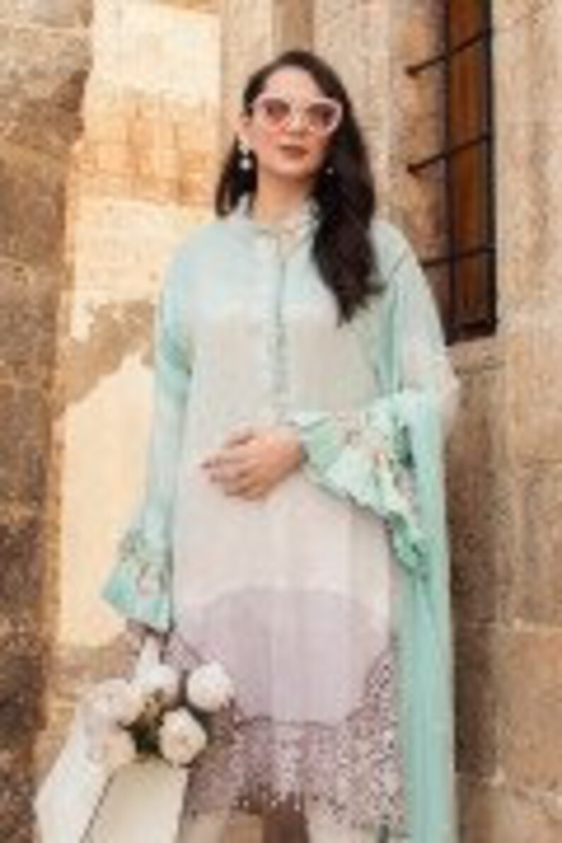 Puede incluir: Una t&uacute;nica de manga larga azul claro y blanca con un dobladillo con volantes y mangas con volantes. La t&uacute;nica tiene un frente abotonado y un estampado floral. La modelo lleva pantalones blancos y sostiene un ramo de flores blancas.