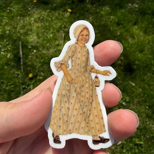 May include: A sticker featuring a smiling girl in a long, patterned dress and bonnet, holding a lantern. The sticker has a white border and is set against a background of green grass.